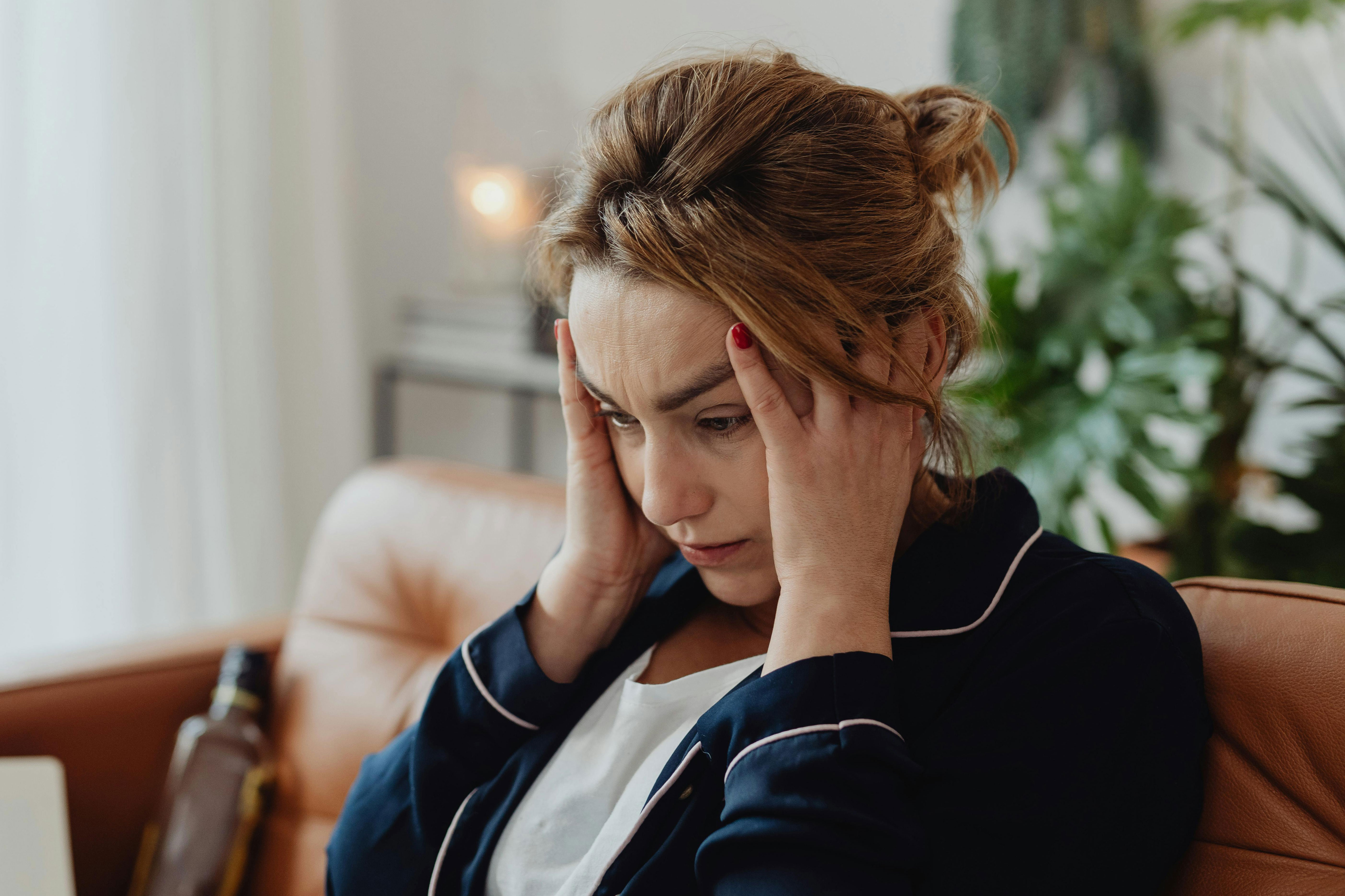 A worried mom sits on a couch with hands on temples, pondering her son's cat allergy.