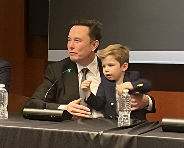 Man in a suit holding a child at a conference table with microphones and water bottles. Man in a suit holding a child at a conference table with microphones and water bottles.