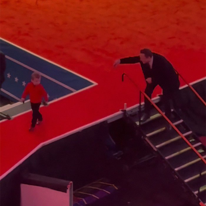Elon Musk on steps reaching towards a young X in a red shirt on a red carpet