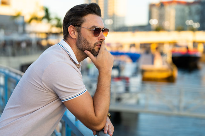 Man in sunglasses pondering near the waterfront under a sunny blue sky, emphasizing concerns about sunscreen and cancer.