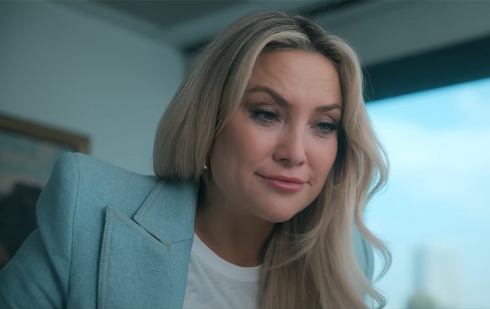 Kate Hudson in a blue blazer looking down with a thoughtful expression, indoors.