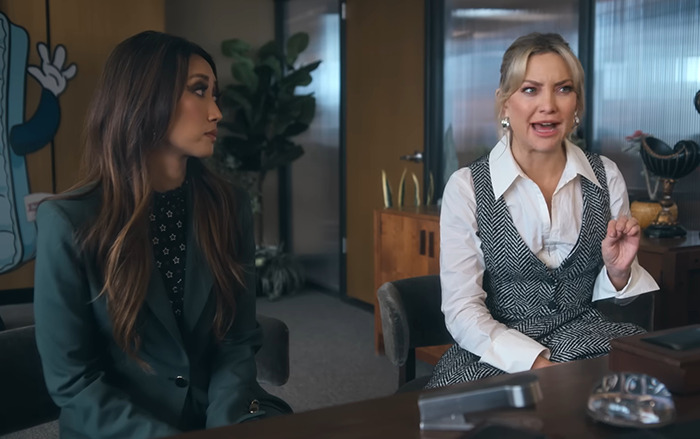 Kate Hudson in an office setting wearing patterned dress and white blouse, engaged in conversation.