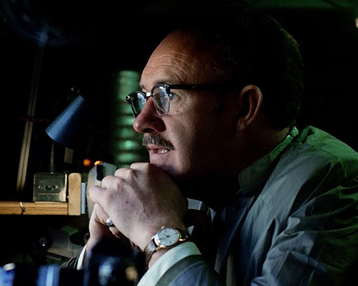 Gene Hackman in a movie scene sitting thoughtfully at a desk, wearing glasses and a watch.