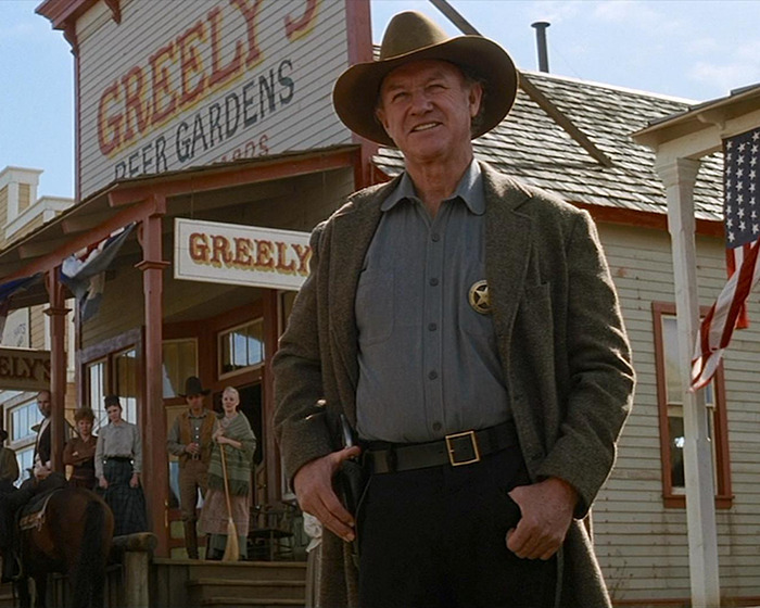 Gene Hackman in a movie scene wearing Western attire standing in front of Greeley's, with onlookers and American flags.