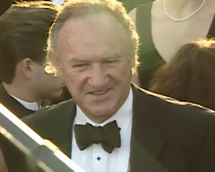 Gene Hackman smiling in a tuxedo at a formal event surrounded by people.