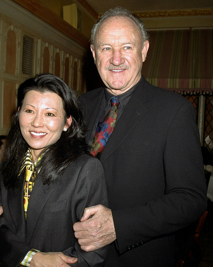 Gene Hackman and Betsy Arakawa smiling together, both wearing black suits indoors.