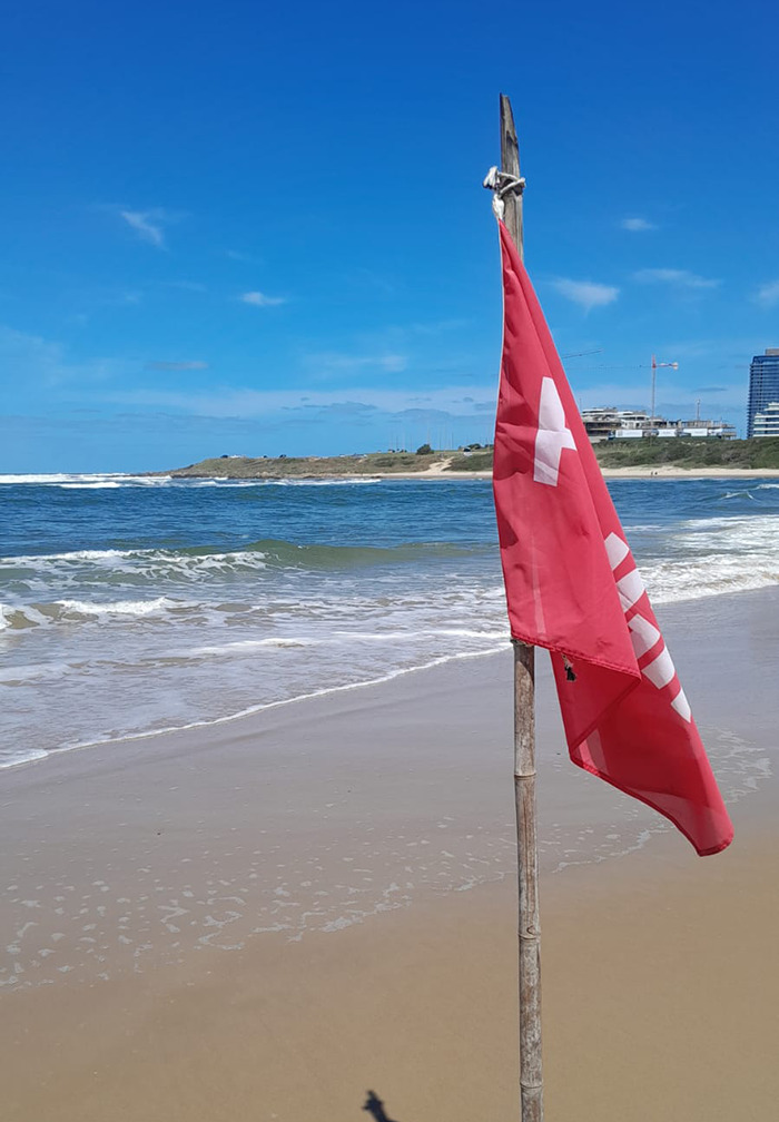 Beach scene with a red lifeguard flag on a pole near the shoreline under a clear blue sky.