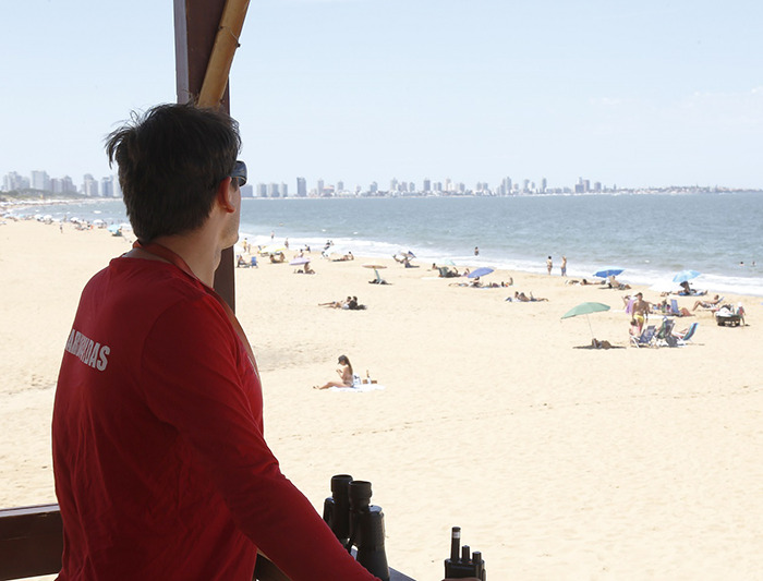 Lifeguard in red shirt overlooking a sunny beach with city skyline in the distance on a clear day.