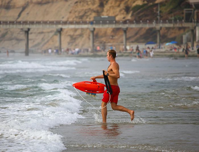 A lifeguard in red shorts holding a lifesaver, running into the ocean.