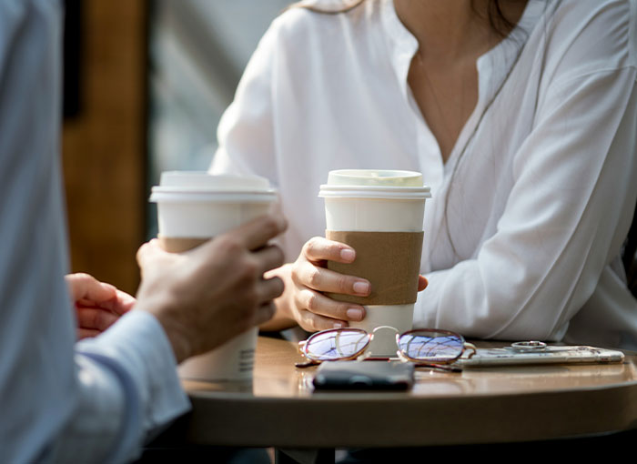 Two colleagues holding coffee cups at a table, one in a white shirt and another partially visible.