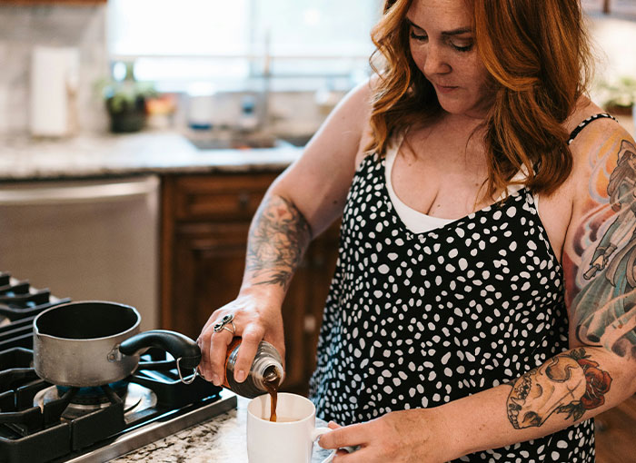 A woman with tattoos pours coffee into a mug in a kitchen setting.