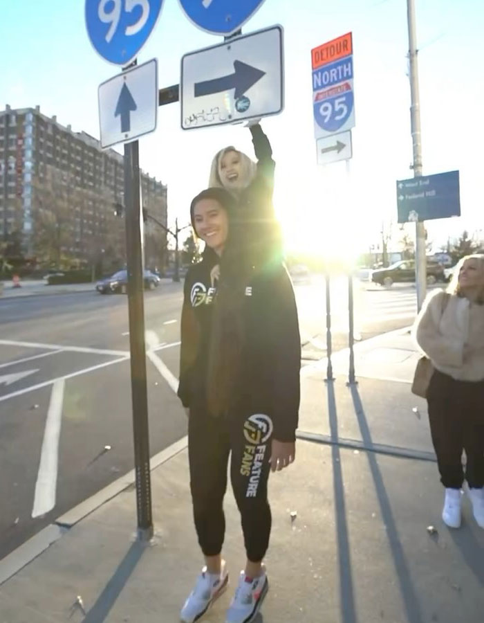 Two people in streetwear under a road sign with the sun setting behind them, featuring a "peculiar" appearance.