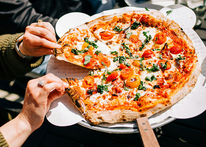 Hands picking up a slice of pizza topped with tomatoes and herbs, a food that people started loving when they grew up.