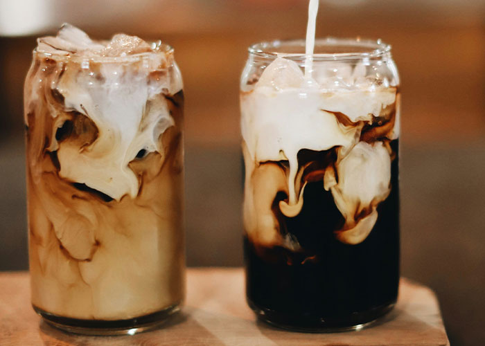 Two glasses of iced coffee with milk swirling, showcasing a food people started loving when they grew up.