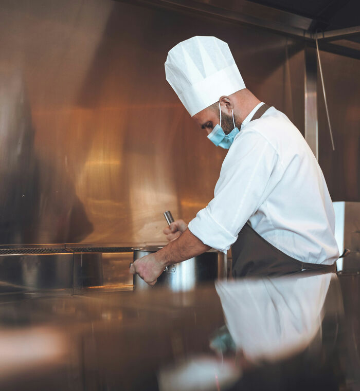 Chef in kitchen, wearing a mask and toque, preparing food; illustrates power dynamics and possible workplace revenge scenario.