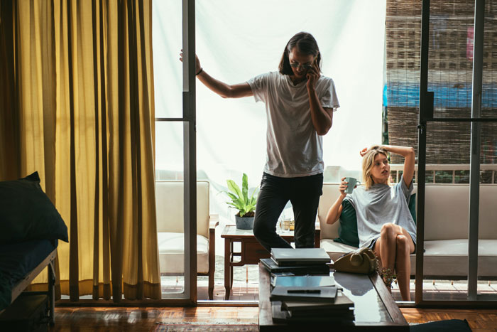 Two people indoors, one standing by the door on the phone, the other sitting with a drink; casual hangout scene. Two people indoors, one standing by the door on the phone, the other sitting with a drink; casual hangout scene.