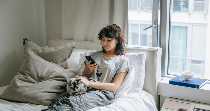 Woman on bed using phone with a cat beside her, looking relaxed and thoughtful.