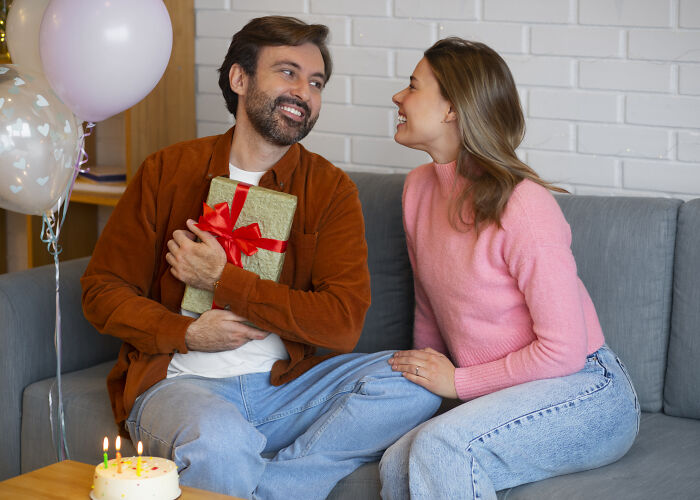 Man receiving a gift from a woman on a couch, surrounded by balloons and a cake, symbolizing love and appreciation.