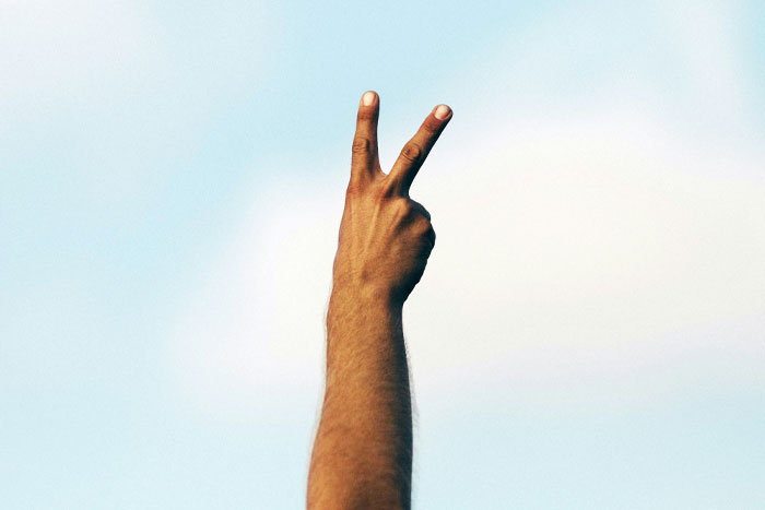 Hand making a peace sign against a blue sky, suggesting funny email sign-offs.