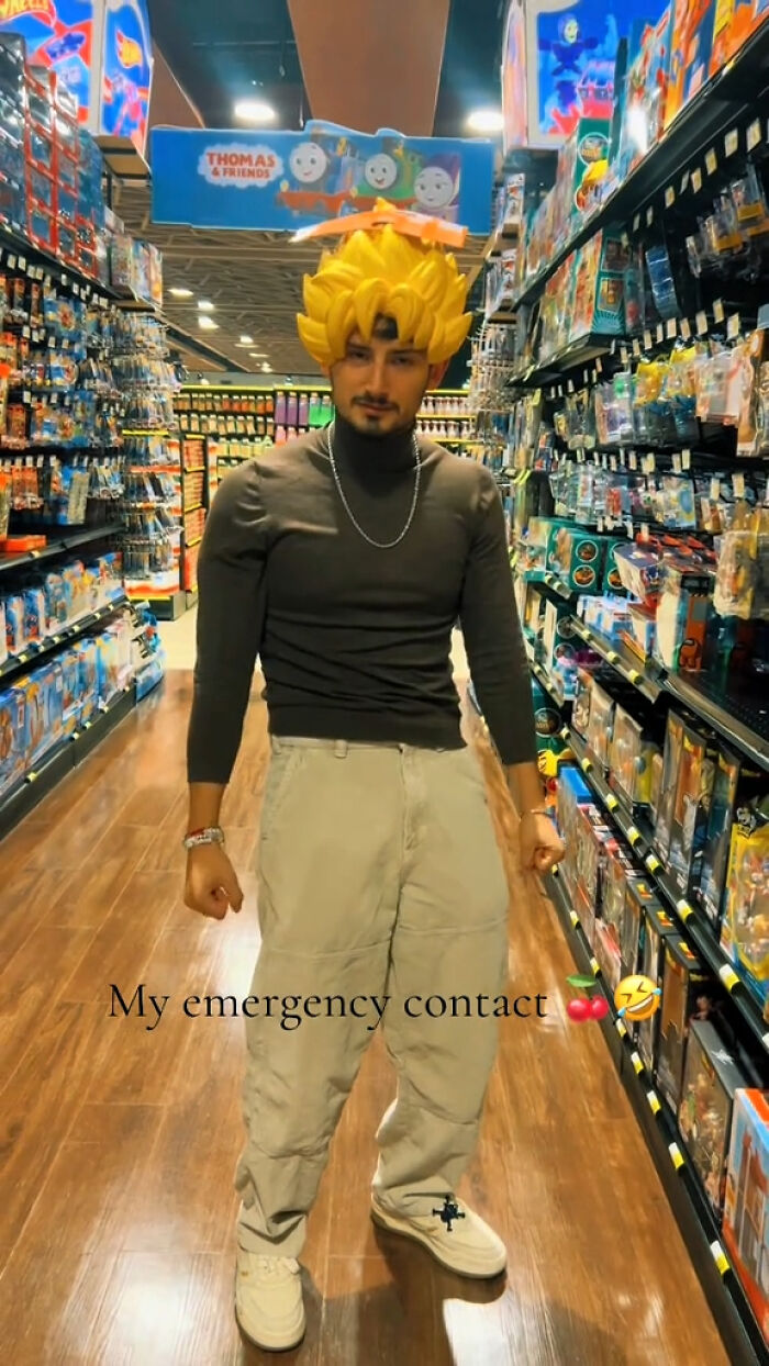 Man in toy aisle wearing a yellow wig and casual clothes, text overlay says "My emergency contact" with emojis.