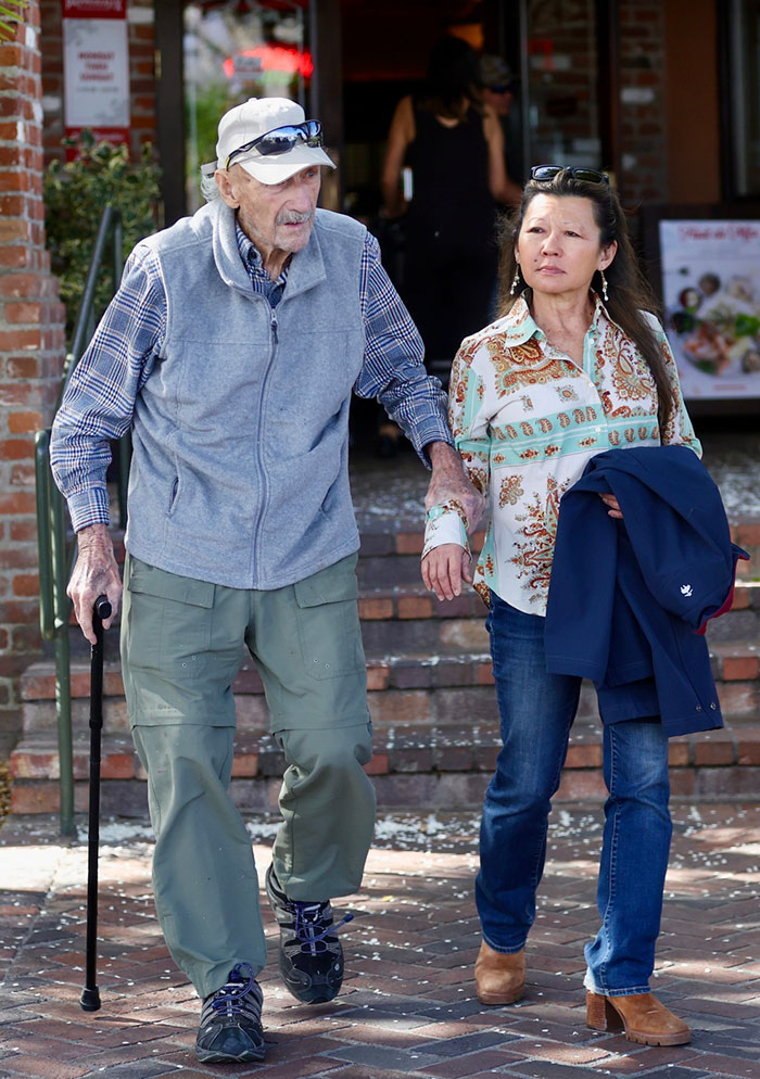 An elderly man and a woman walking together outdoors, with the man using a cane for support.