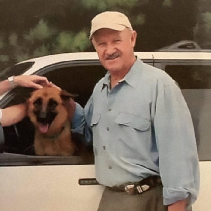 Gene Hackman in a blue shirt and cap stands by a car with a dog inside, smiling outdoors.