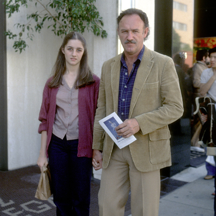 Gene Hackman and daughter holding hands on a sidewalk, near a potted plant, outside a building.