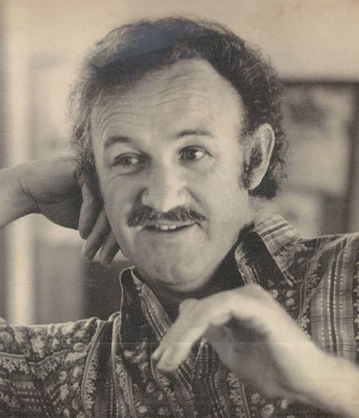 An older image of Gene Hackman smiling in a casual shirt, gesturing with a relaxed demeanor.