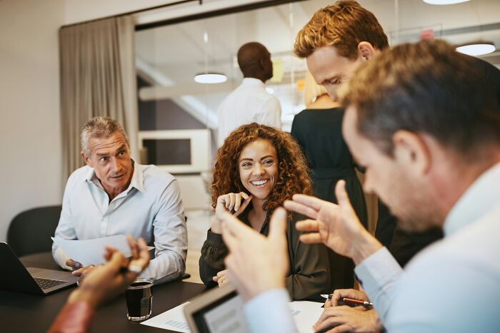 People in a lively office meeting, a woman smiling while others discuss around her, illustrating unintended offensive remarks.