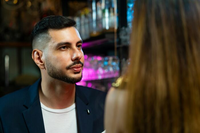 A man in a suit listens to a woman in a bar, highlighting unintentional offensive remarks towards women.