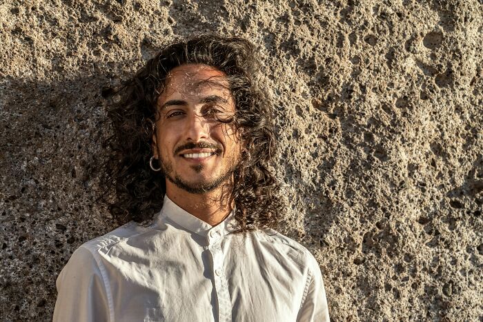 Man with curly hair and prominent nose, wearing a white shirt, smiling against a textured stone wall.