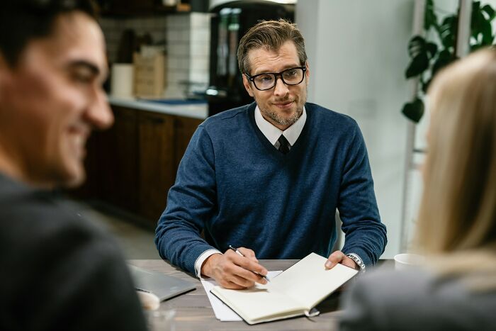 Man in an office, wearing glasses and a sweater, writing in a notebook.