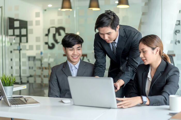Three professionals in a modern office collaborate at a laptop, discussing women’s experiences with unintentional offensive remarks.