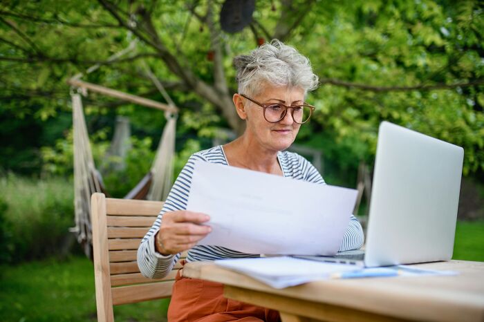 Senior woman working outdoors on a laptop, reviewing documents, representing professions that employ millions.