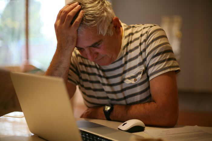 Man in striped shirt, holding his head while looking at a laptop, illustrating adulthood challenges.