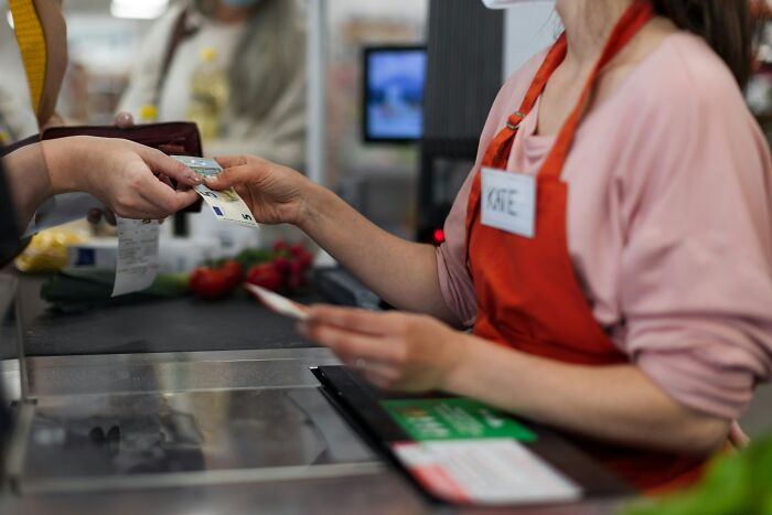 Cashier in a red apron accepting money from a customer at a busy checkout counter in a workplace moment.