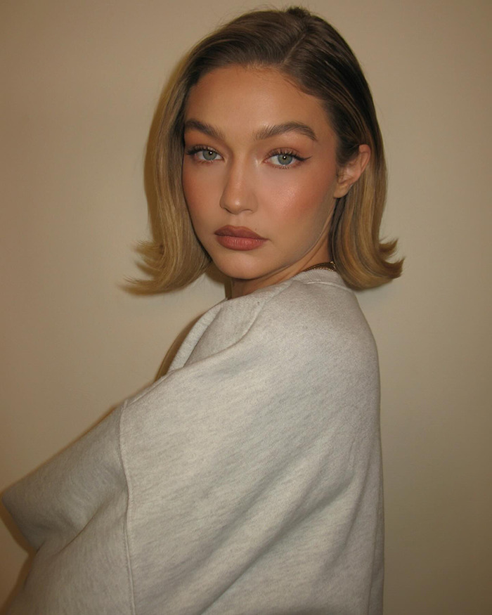 Gigi Hadid in a white sweater, looking over her shoulder with a neutral background.