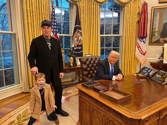 Elon Musk in a suit with son in a coat standing next to President Donald Trump who's seated at a large desk in an office with flags behind. Elon Musk in a suit with son in a coat standing next to President Donald Trump who's seated at a large desk in an office with flags behind.