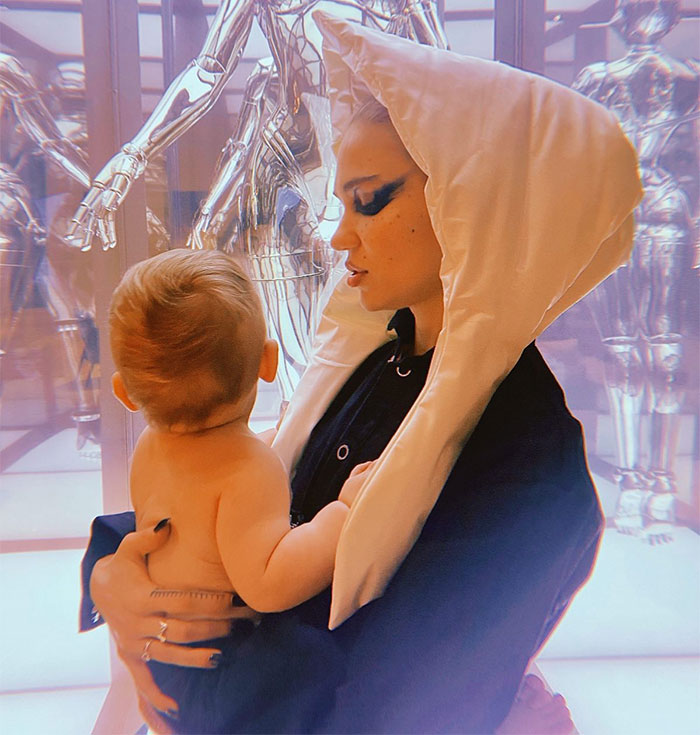Grimes holding a baby, dressed in a unique outfit, against a futuristic background.