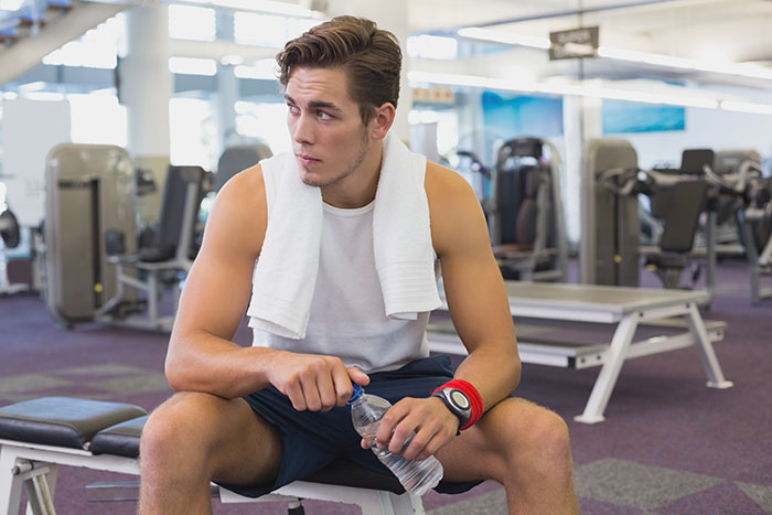 Man with towel in gym sitting, looking contemplative, holding a water bottle. Fitness equipment in background. Man with towel in gym sitting, looking contemplative, holding a water bottle. Fitness equipment in background.