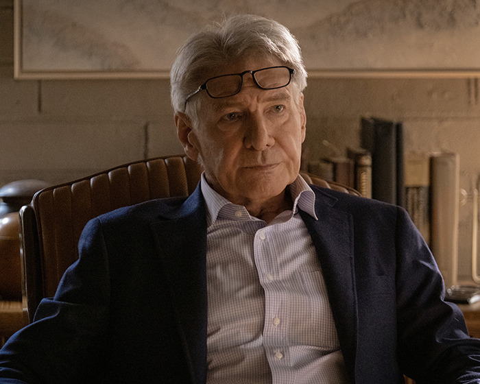 Harrison Ford seated with glasses atop head, wearing a suit and light shirt, thoughtful expression against bookshelf backdrop in a scene from "Shrinking.