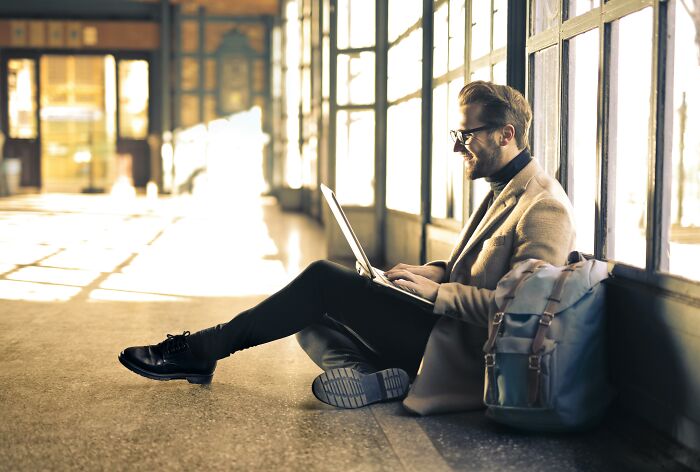 Man sitting on the floor with a laptop, wearing glasses and a coat, sunlight streaming in. 