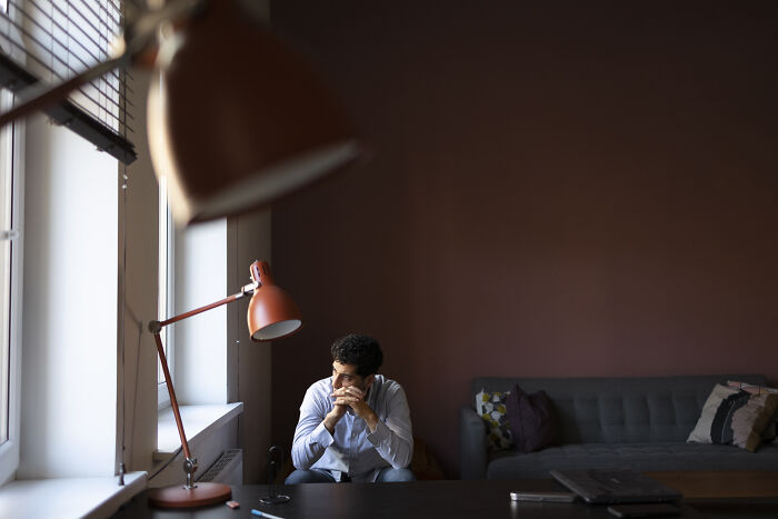 A man sits alone, looking pensive, in a dimly lit room, symbolizing a heartbroken friend feeling like a house leech.