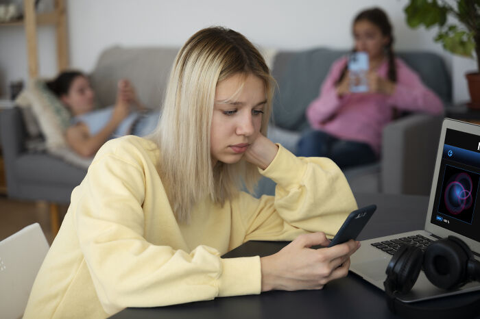 Young woman in a yellow sweater looking at her phone, with two people on a sofa using devices in the background.