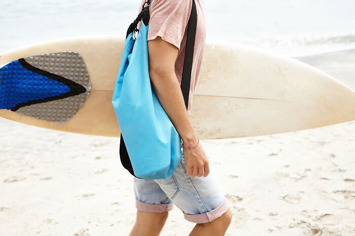 Person at the beach with a blue travel essentials bag, carrying a surfboard.