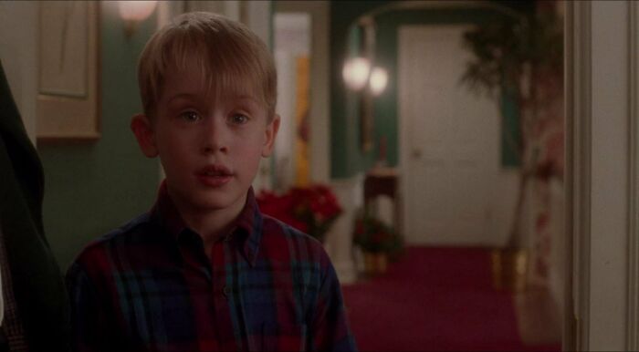 Young actor in a 90s film scene, wearing a plaid shirt, standing in a warmly decorated hallway.