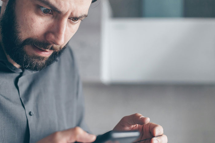 Man looking shocked at phone, wearing a grey shirt, representing a husband's reaction to a wife's actions.