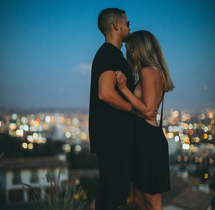 A couple embracing on a rooftop at night, with city lights in the background, depicting fantasy romance tropes.