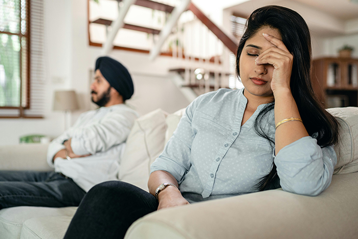 Woman looking stressed on couch; upset husband in the background.