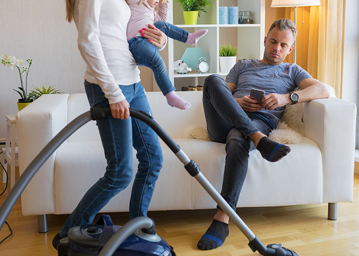 Husband on couch using phone while wife vacuums holding child, highlighting "unhelpful" dynamic.